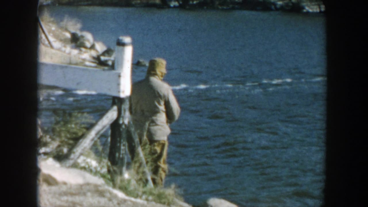 Man seen from behind fishing beside blue river water and rocks.