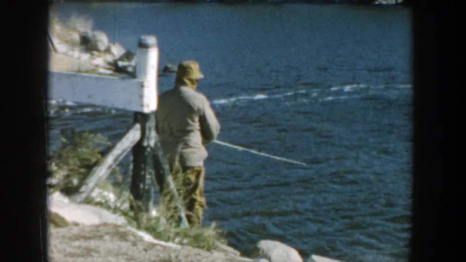 A man in a hat fishes at the edge of a Wisconsin river, with his line visible across the rippling water.