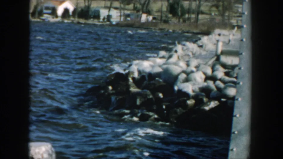 The view shifts to the rocky river edge and moving water, with a distant shoreline visible in the background.