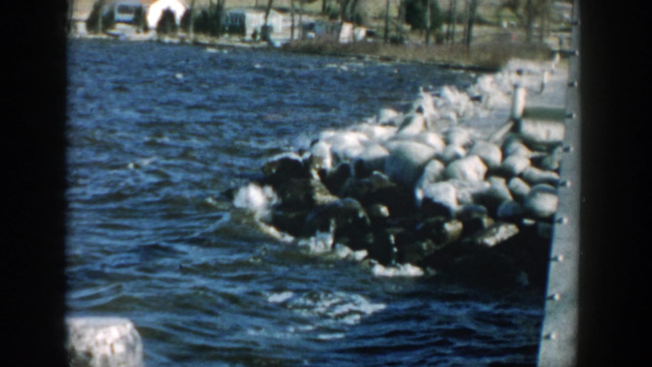Choppy blue water hits large rocks beside a metal structure, with houses and bare trees in the background.