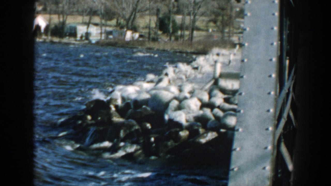Water splashes around dark and pale shoreline rocks near a riveted metal structure.
