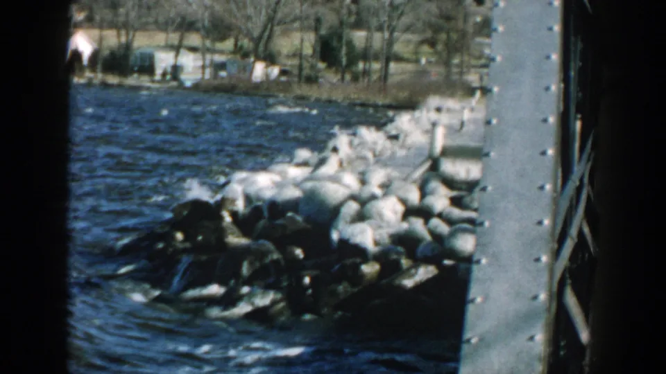 Choppy Wisconsin lake water breaks over large shoreline stones beside a metal structure in this 1957 8mm frame.