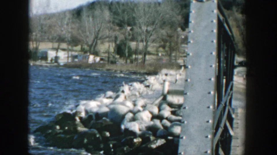 Wisconsin lake shoreline in 1957, showing waves at a rocky breakwater framed by a riveted metal bridge or pier element.