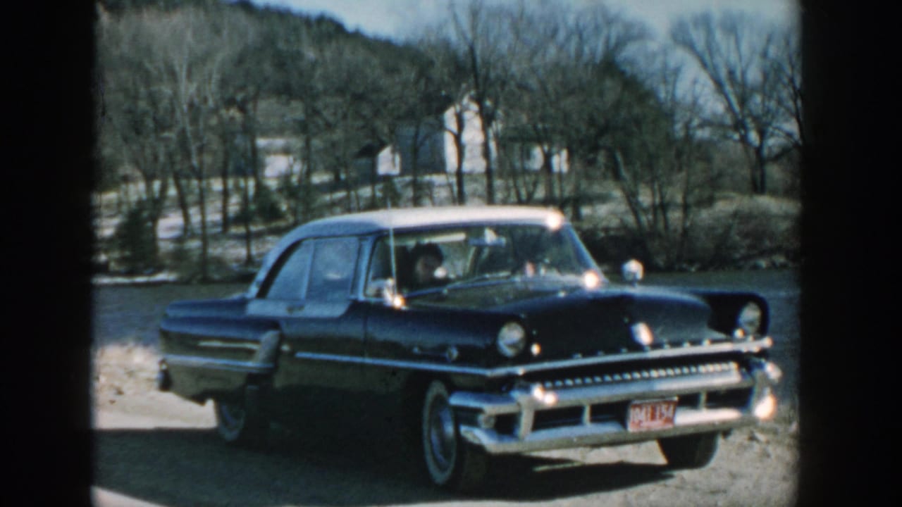 Black sedan with white roof parked on gravel near bare trees and rural buildings.