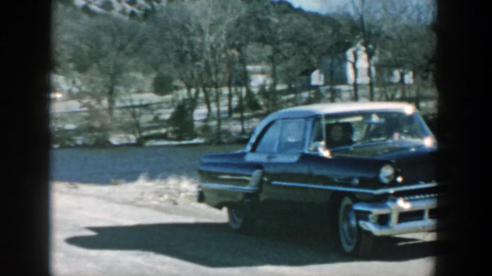A mid-century black sedan with white roof is shown in partial profile on gravel, with bare trees and rural Wisconsin buildings in the background.