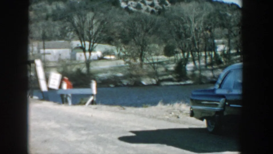 The car moves partly out of frame or is framed at the edge, revealing more of the rural Wisconsin roadside, bare trees, and distant structures.