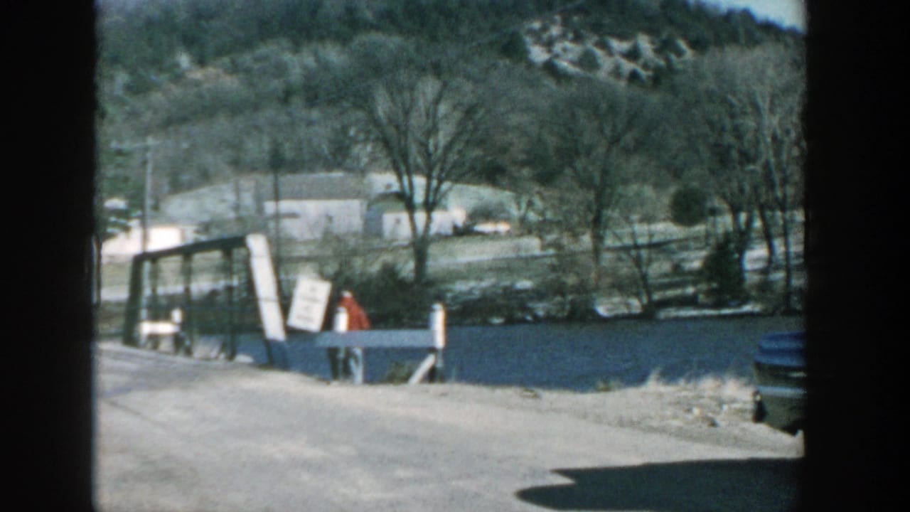 1957 Wisconsin 8mm frame showing a rural bridge or crossing beside water, a distant person in red, bare trees, and low wooded hills.