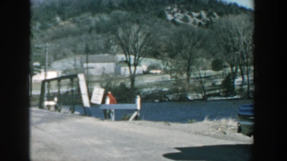 1957 Wisconsin 8mm frame showing a rural bridge or crossing beside water, a distant person in red, bare trees, and low wooded hills.