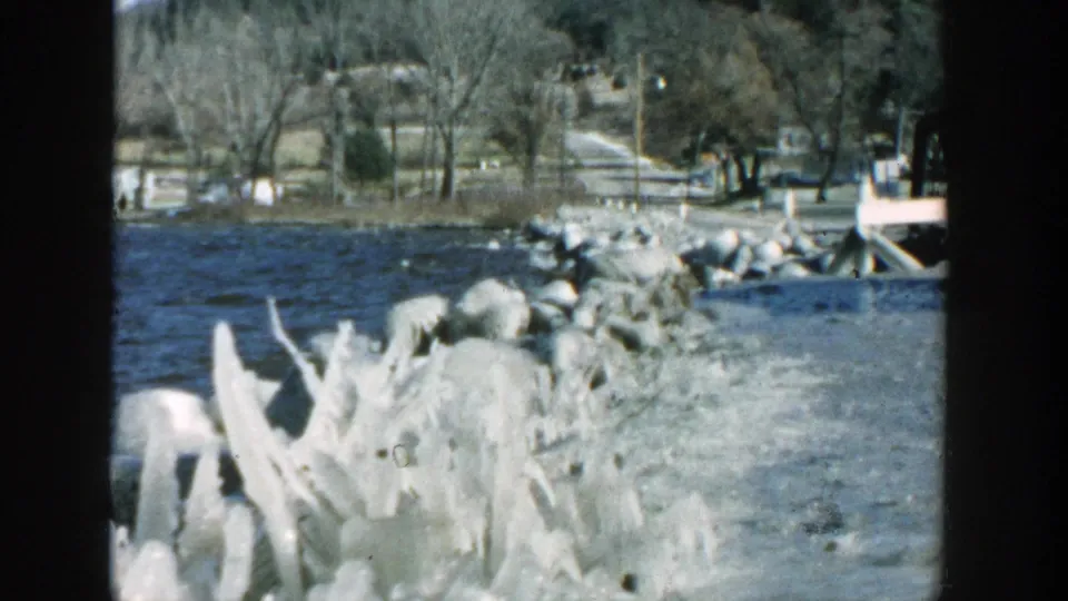 1957 Wisconsin 8mm frame looking across a rocky lakeside or riverside edge toward a rural road, bare trees, and hillside.
