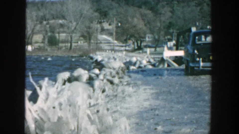 1957 Wisconsin home-movie frame with water, pale shoreline rocks, a rural road, bare trees, and a dark vehicle at the right edge.