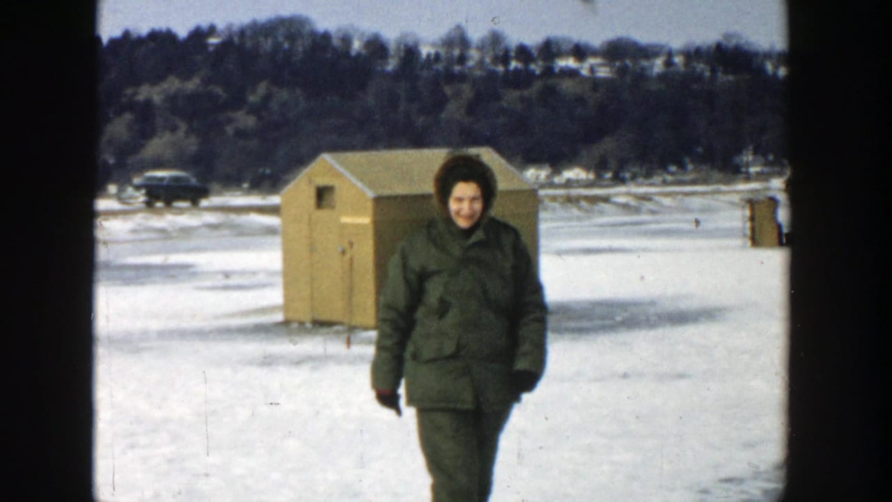 In 1957 Wisconsin home-movie footage, a woman in a green winter coat walks across a frozen lake toward the camera near an ice-fishing shed.