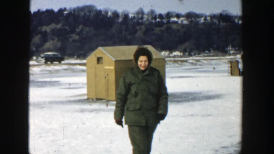 In 1957 Wisconsin home-movie footage, a woman in a green winter coat walks across a frozen lake toward the camera near an ice-fishing shed.