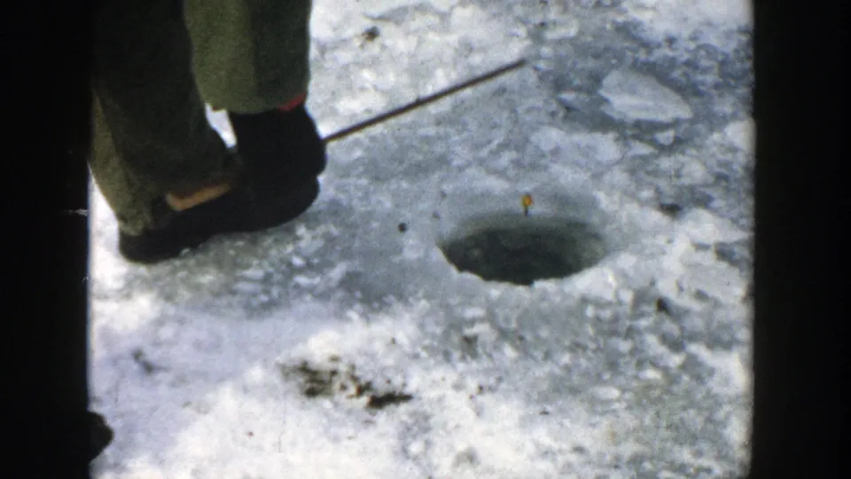 A close view shows the woman's boots and winter clothing beside a round ice-fishing hole cut into the frozen lake surface.
