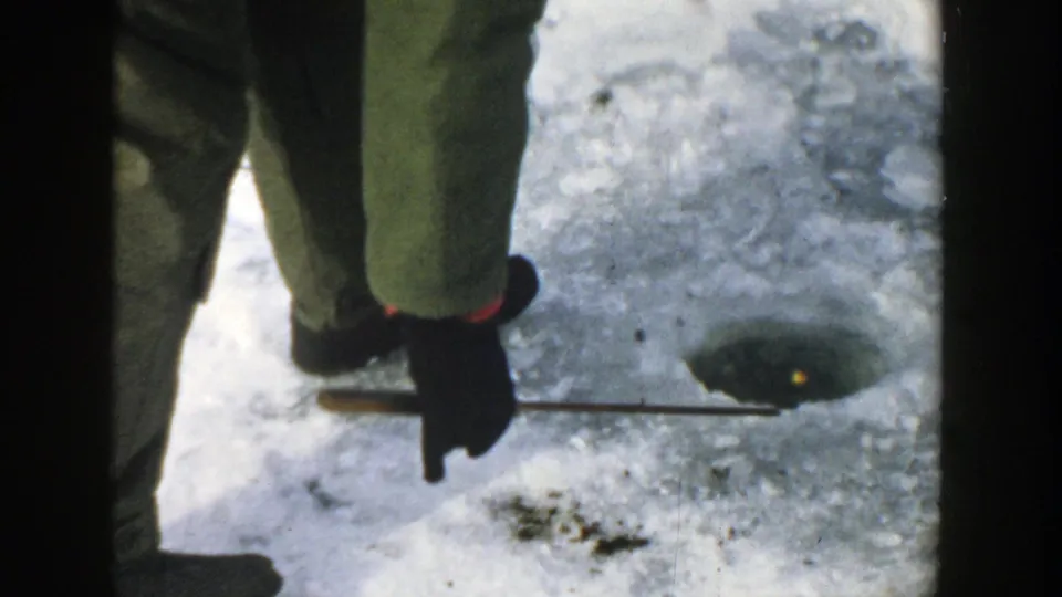 The woman lowers a short rod or line toward an ice-fishing hole, with her gloved hand and boots visible on the frozen lake.