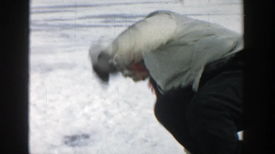 In 1957 Wisconsin home-movie footage, a woman in a white coat bends low on the snow-covered ice of a frozen lake.