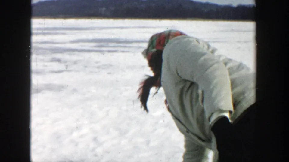 A woman in a white winter coat and red plaid headscarf leans forward on a snow-covered frozen Wisconsin lake.