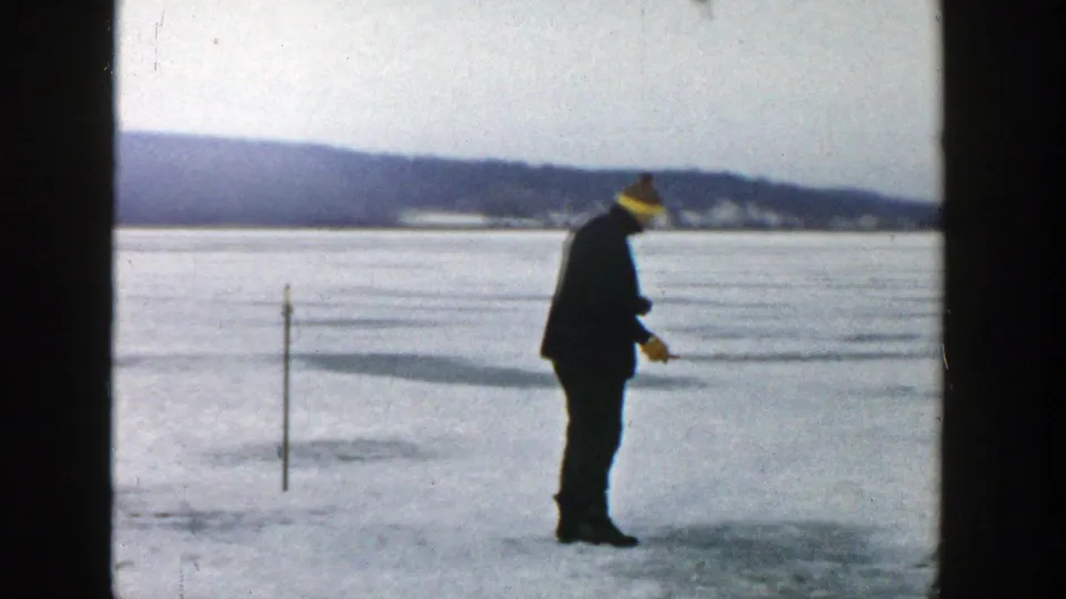 A man in a dark coat and yellow hat stands on the frozen lake near a small pole, holding a tool toward the ice.