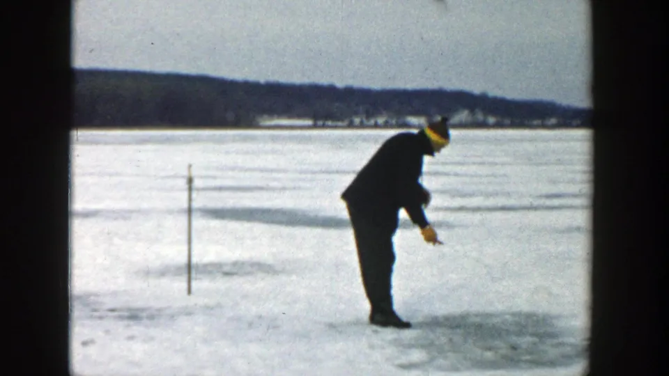 The man bends over the ice with a small tool, with a marker pole and distant Wisconsin shoreline behind him.