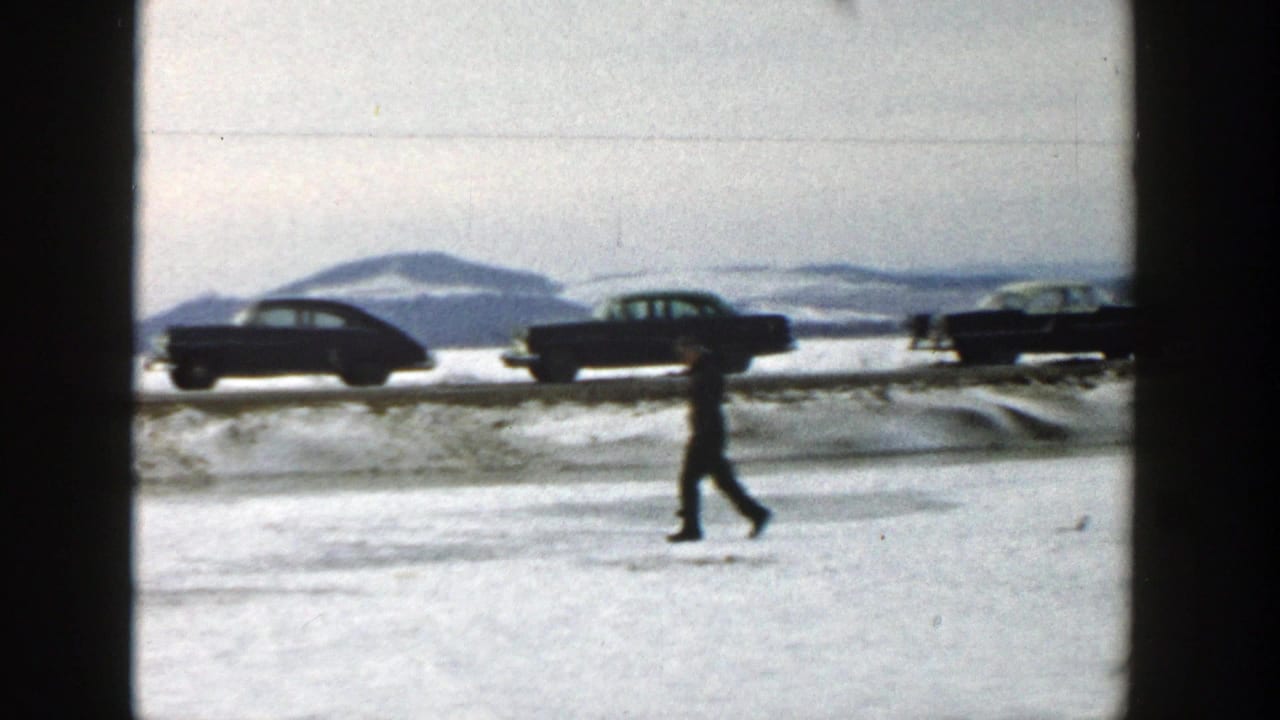 Man walking across snow with cars passing on a road behind him.