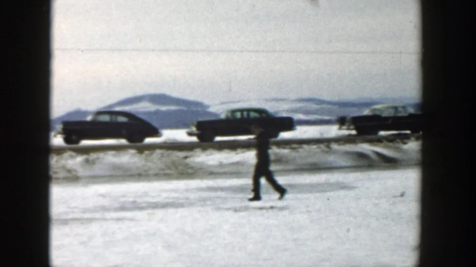 In a 1957 Wisconsin winter home-movie frame, a man crosses a snowy field while mid-century cars move along a road beyond him.