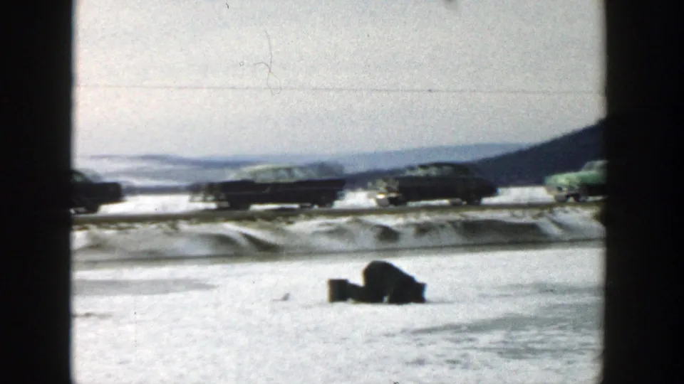 A snowy Wisconsin roadside scene shows several 1950s-era cars moving past, with a dark object or person low in the snow at right.