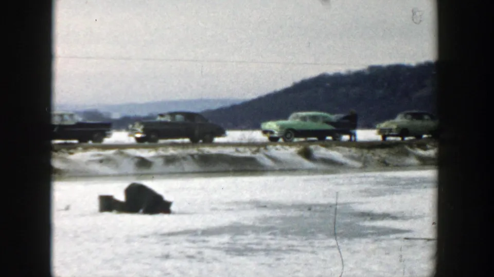 A 1957 Wisconsin winter frame shows mid-century cars, including a green sedan, traveling along a snow-lined road with hills beyond.