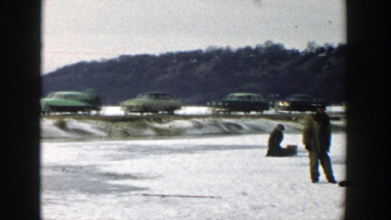 Two men on a frozen lake with cars passing along a snowy road behind them.
