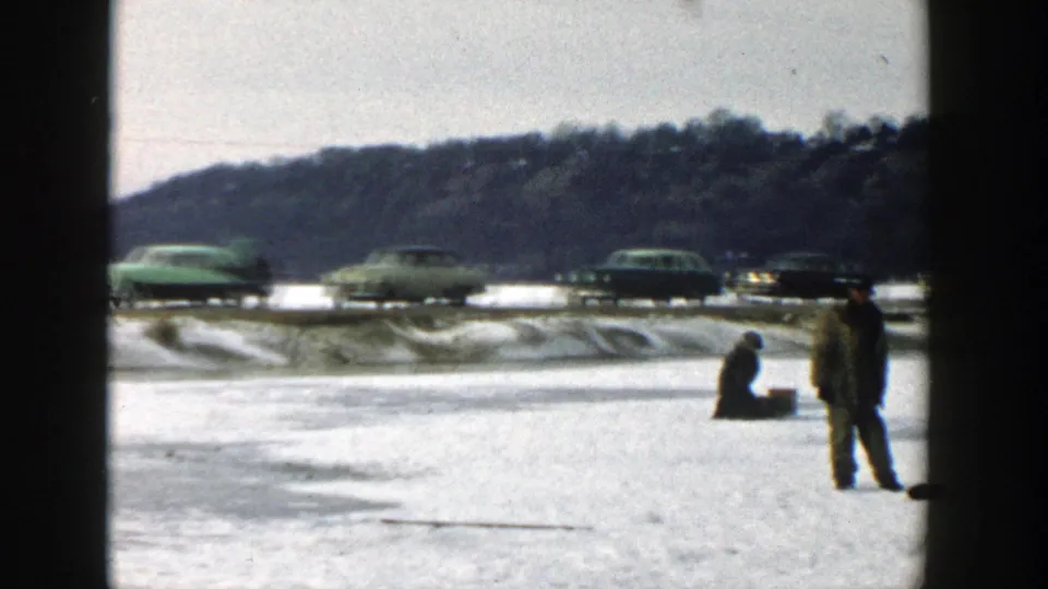 Wisconsin 1957 home-movie frame showing two adult men ice fishing on a frozen lake with mid-century cars in the background.