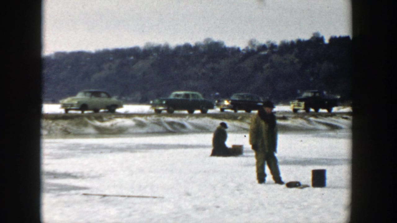 Standing man and seated man on snowy lake ice with cars and gear nearby.