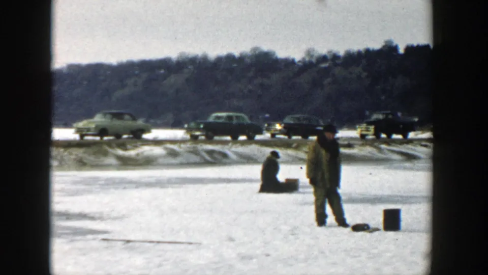 Wisconsin 1957 frame of a standing man near fishing gear and a seated man on frozen lake ice, with cars beyond the shoreline.
