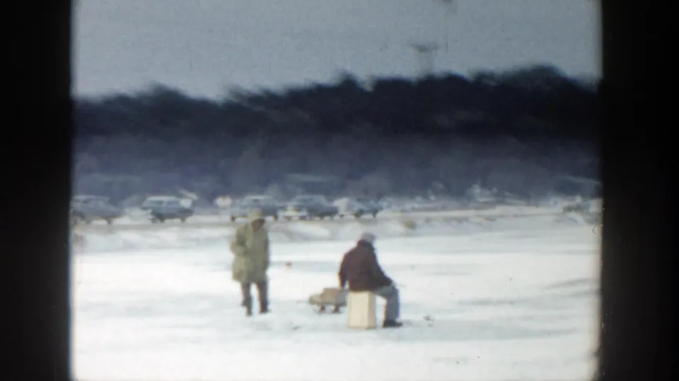Distant Wisconsin 1957 home-movie view of two men ice fishing on a snowy frozen lake with traffic and trees in the background.