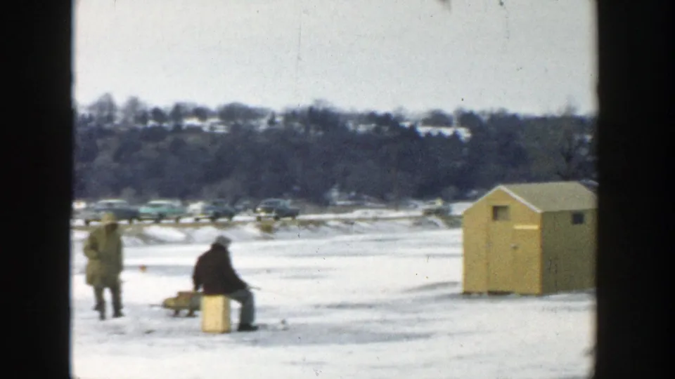 Wisconsin 1957 frame showing two men on lake ice, a seated fisher, passing cars, and a small yellow ice-fishing hut nearby.