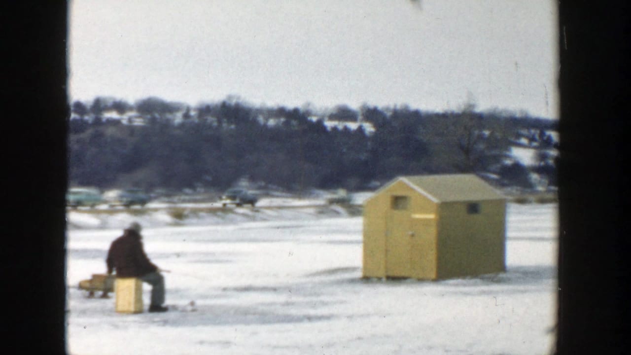 1957 Wisconsin 8mm frame showing a seated figure beside a crate and yellow ice-fishing hut on snow-covered lake ice.