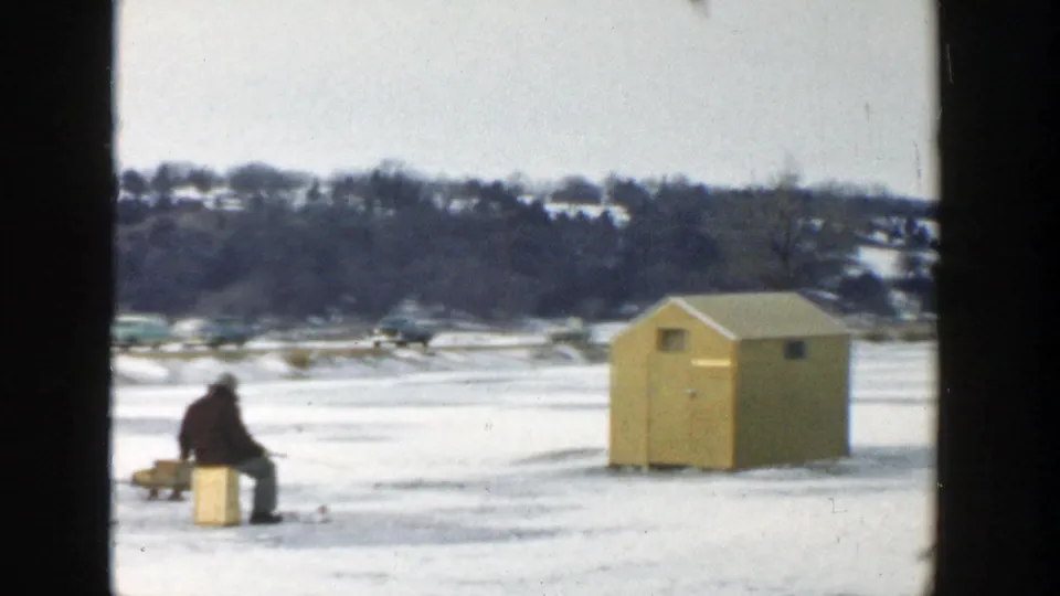 1957 Wisconsin 8mm frame showing a seated figure beside a crate and yellow ice-fishing hut on snow-covered lake ice.