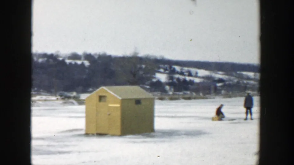 1957 Wisconsin winter frame with a yellow ice-fishing hut and two distant figures on a snow-covered frozen lake.