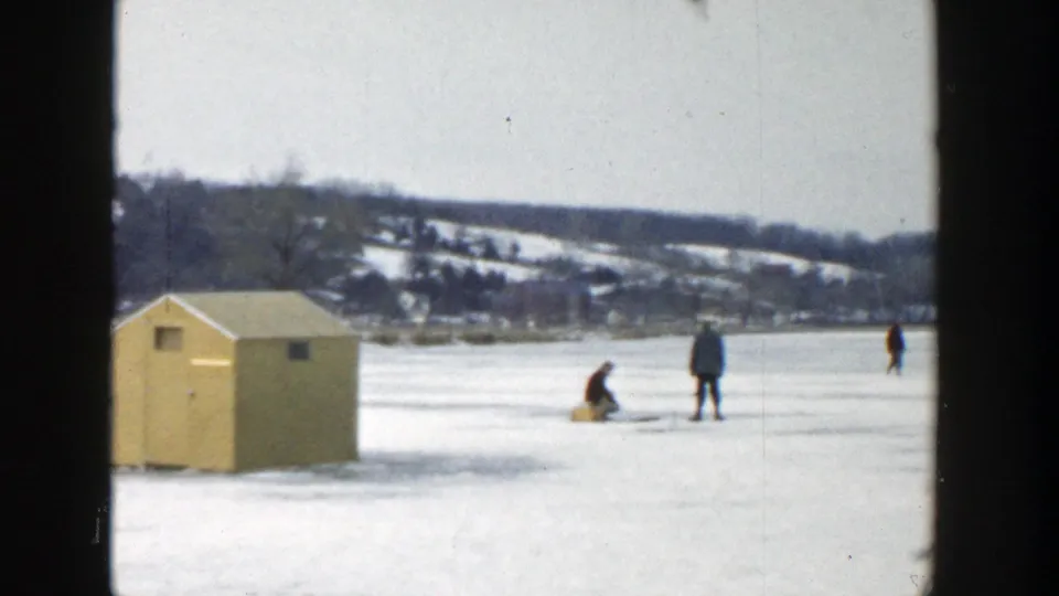 1957 Wisconsin home-movie frame showing a yellow hut and several distant figures walking or fishing on a frozen lake.