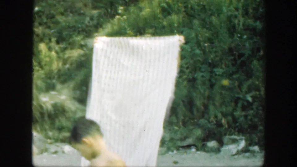 A white textured sheet dominates the 8mm frame while the man bends low at the edge of the outdoor scene.
