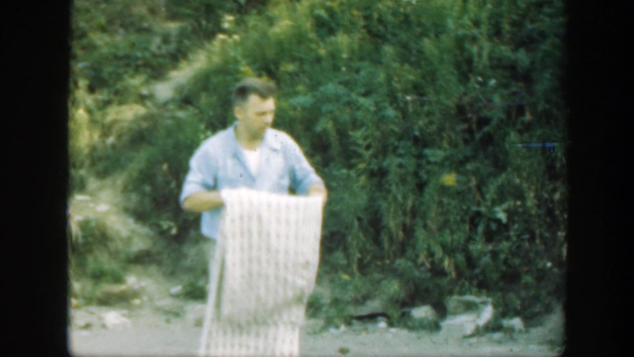 Man in a light shirt holds a pale checkered blanket beside dense green woods.
