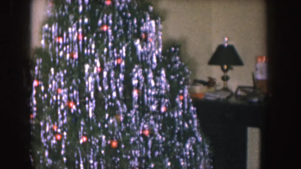 1955 Nebraska home-movie frame showing a Christmas tree covered in silver tinsel and red ornaments beside a table lamp in a private interior.