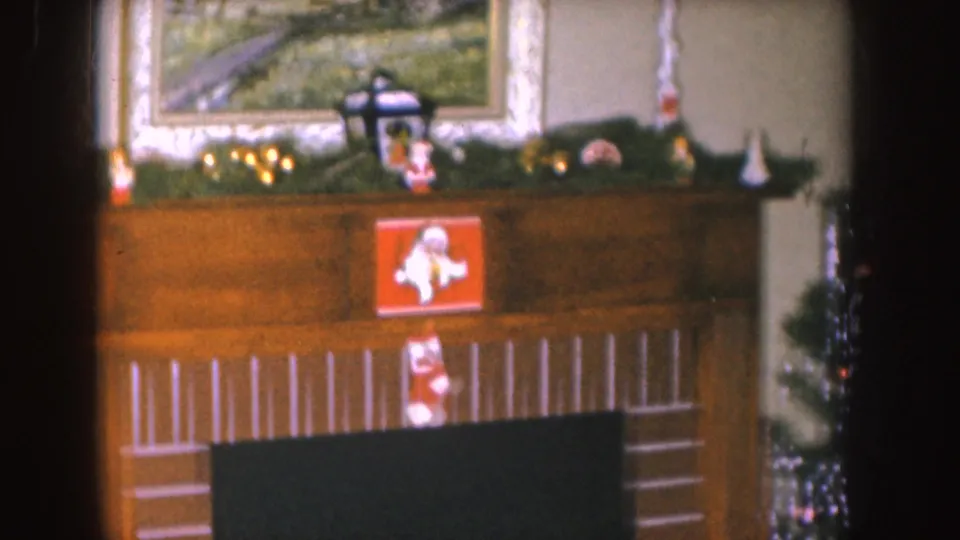 1955 Nebraska home-movie frame of a Christmas mantel display with stockings, greenery, framed wall art, and a tinsel tree partly visible.