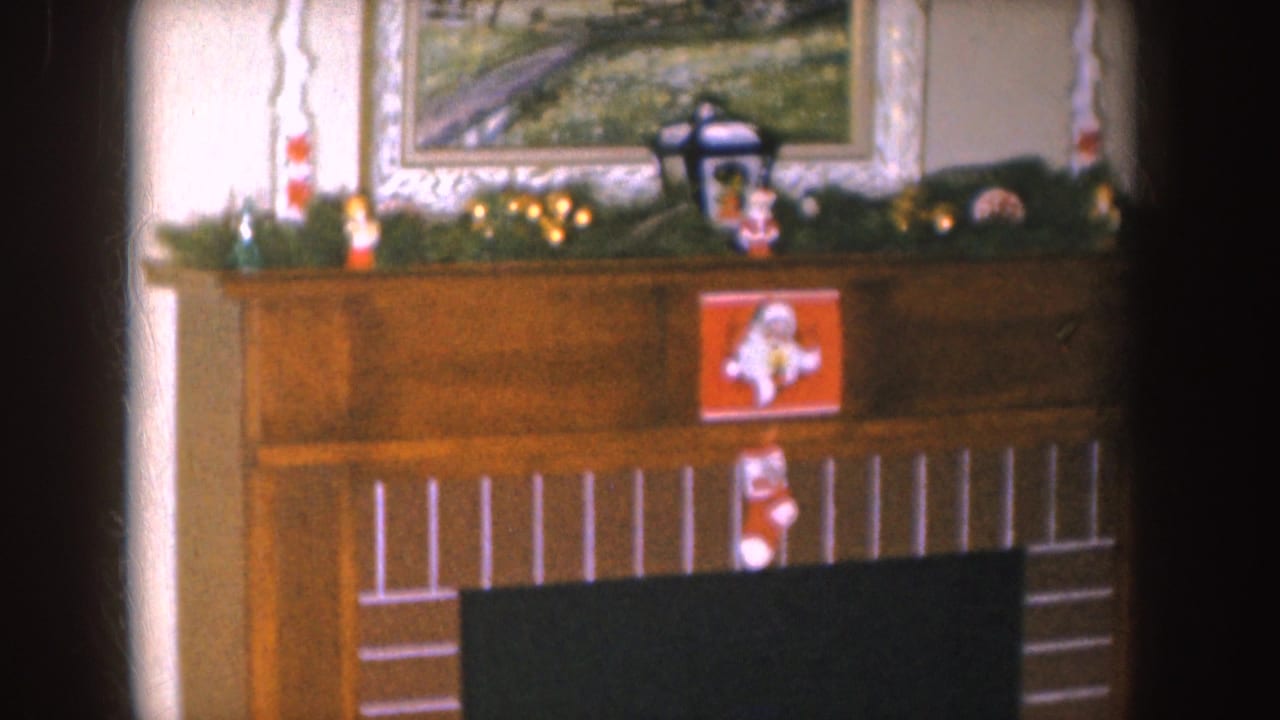 1955 Nebraska home-movie frame showing a decorated fireplace mantel with garland, a Santa card, candles, and a framed landscape painting.
