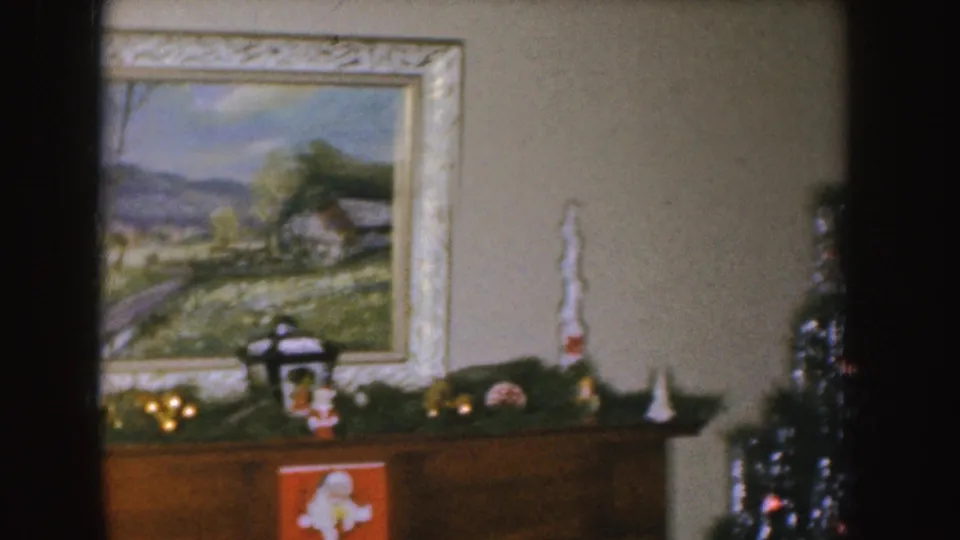 1955 Nebraska frame of a holiday mantel arrangement with garland, small decorations, a framed landscape painting, and part of a Christmas tree.