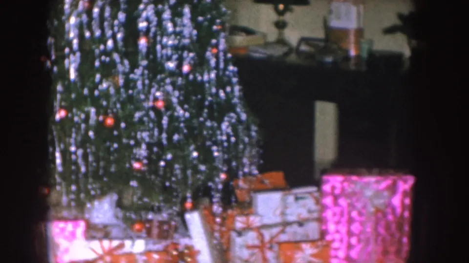 Nebraska 1955 home-movie view of a tinsel Christmas tree beside a mantel, with wrapped gifts clustered below.
