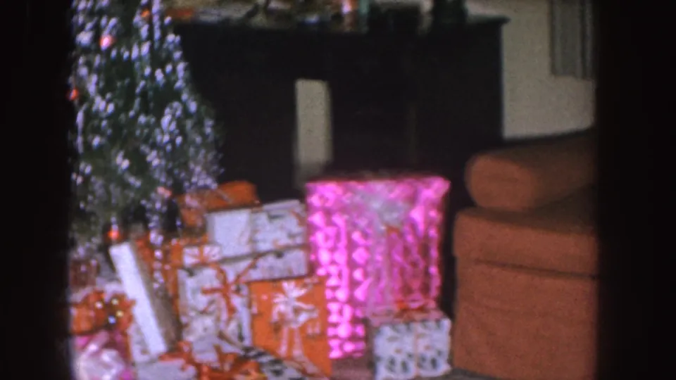 Nebraska 1955 frame showing gifts under a tinsel Christmas tree, a fireplace mantel, and an upholstered chair at right.