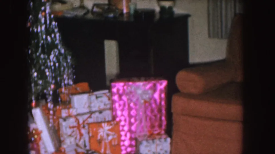 1955 Nebraska home-movie frame showing wrapped presents near a tinsel-covered Christmas tree, with a console and armchair behind them.
