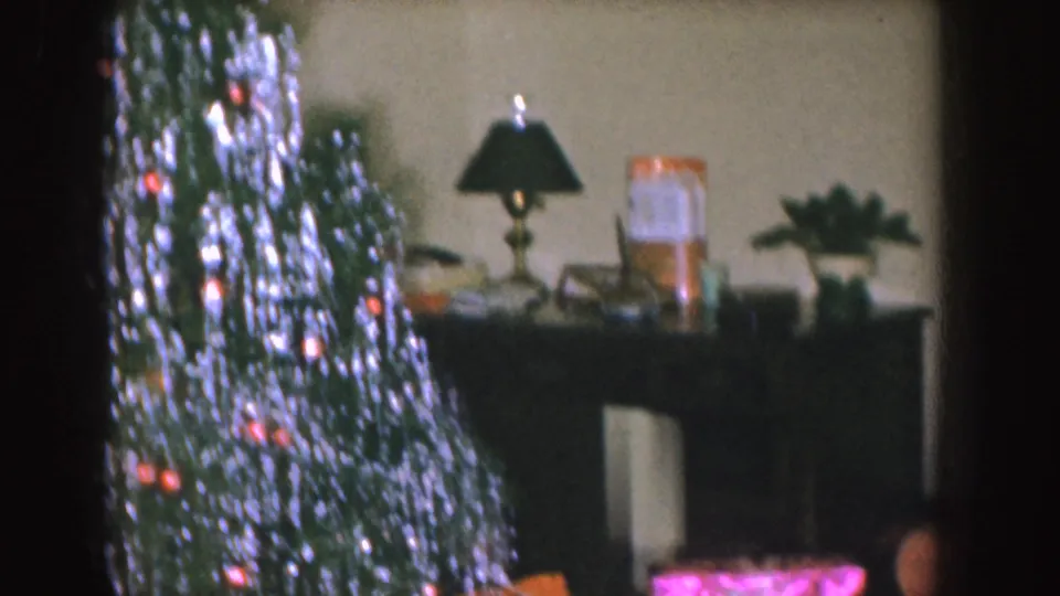 1955 Nebraska home-movie frame focused on the tinsel-covered Christmas tree, wrapped gifts, a console table, lamp, and plant.