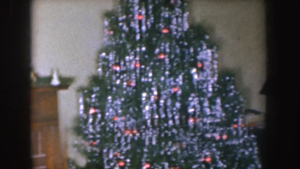 1955 Nebraska home-movie frame showing a decorated Christmas tree with silver tinsel and red lights beside household furniture.