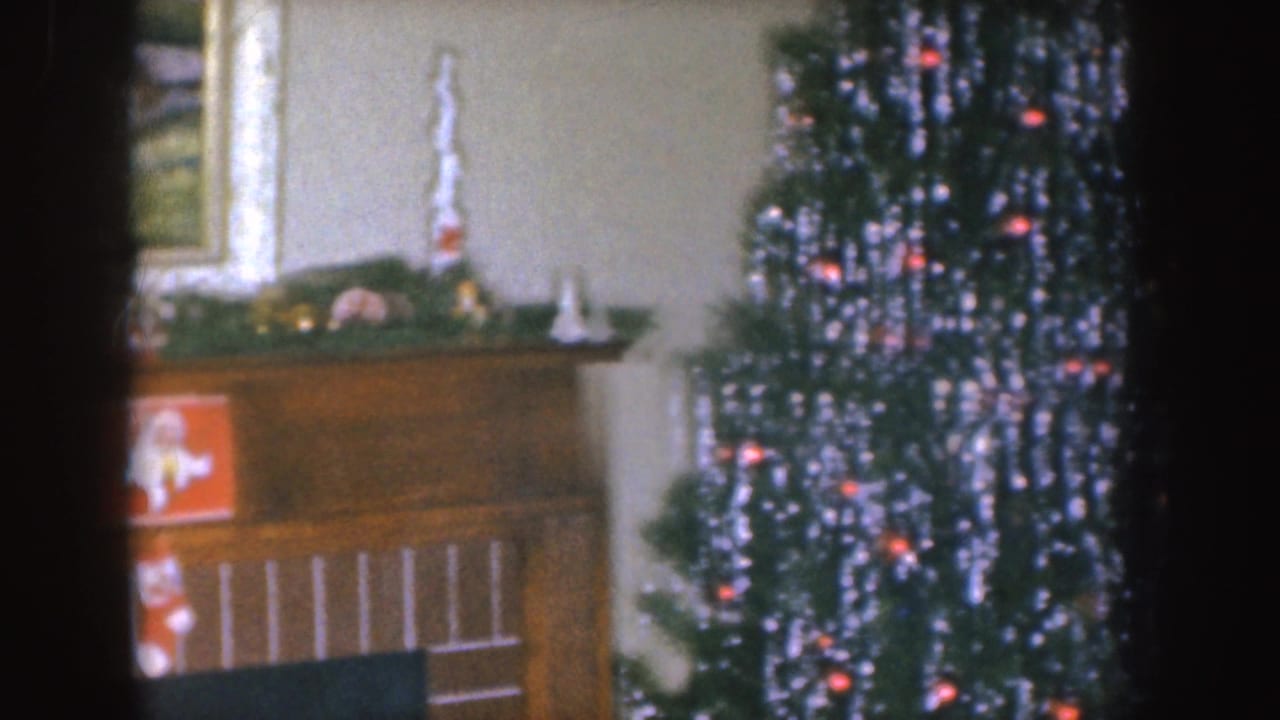 Nebraska 1955 home-movie frame showing a Christmas mantel with garland and a lit tinsel tree in a private interior.