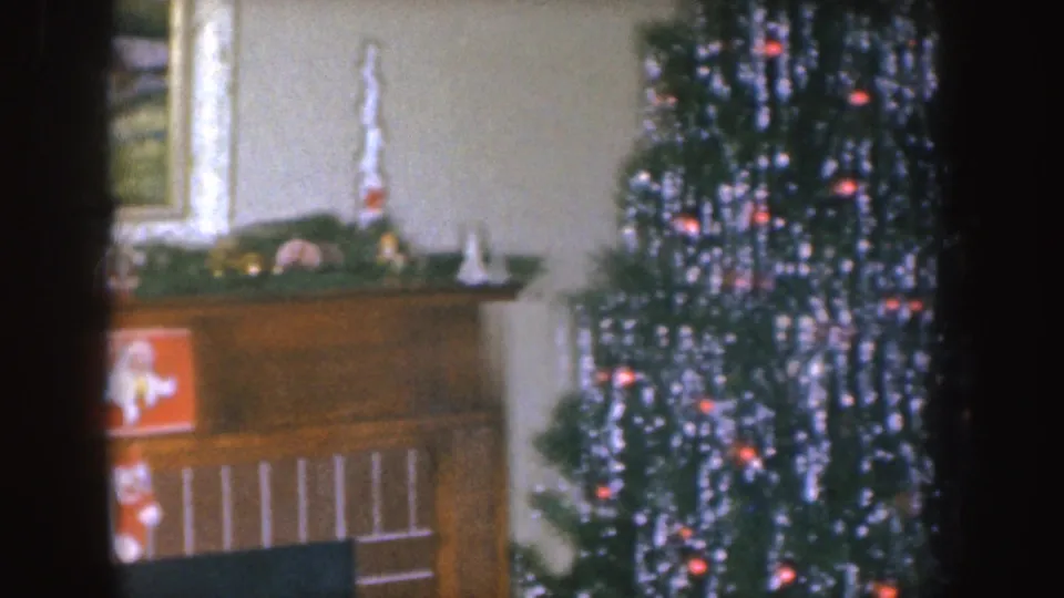 Nebraska 1955 home-movie frame showing a Christmas mantel with garland and a lit tinsel tree in a private interior.
