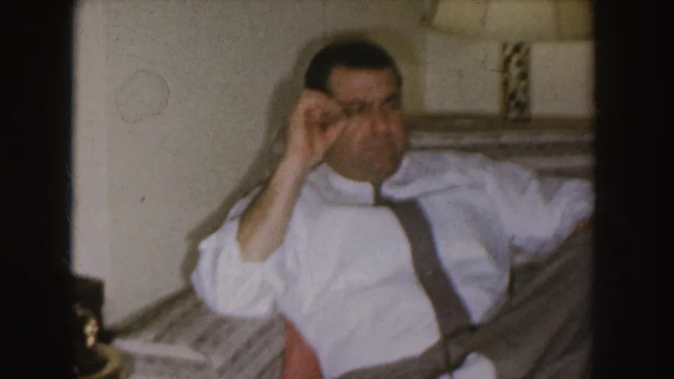 Nebraska 1955 home-movie frame showing a man in a white shirt and suspenders reclining on a sofa during a Christmas gathering.